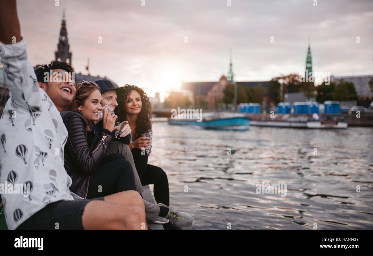 Shot of group of people sitting outdoors on jetty and having fun. Friends hanging out on pier over lake in the city. Stock Photo