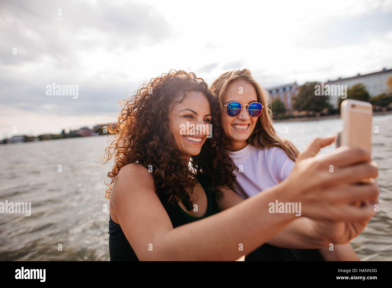 Shot of cheerful teenage girls smiling and taking selfie with smart phone by the lake. Female friends taking self portrait with mobile phone. Stock Photo