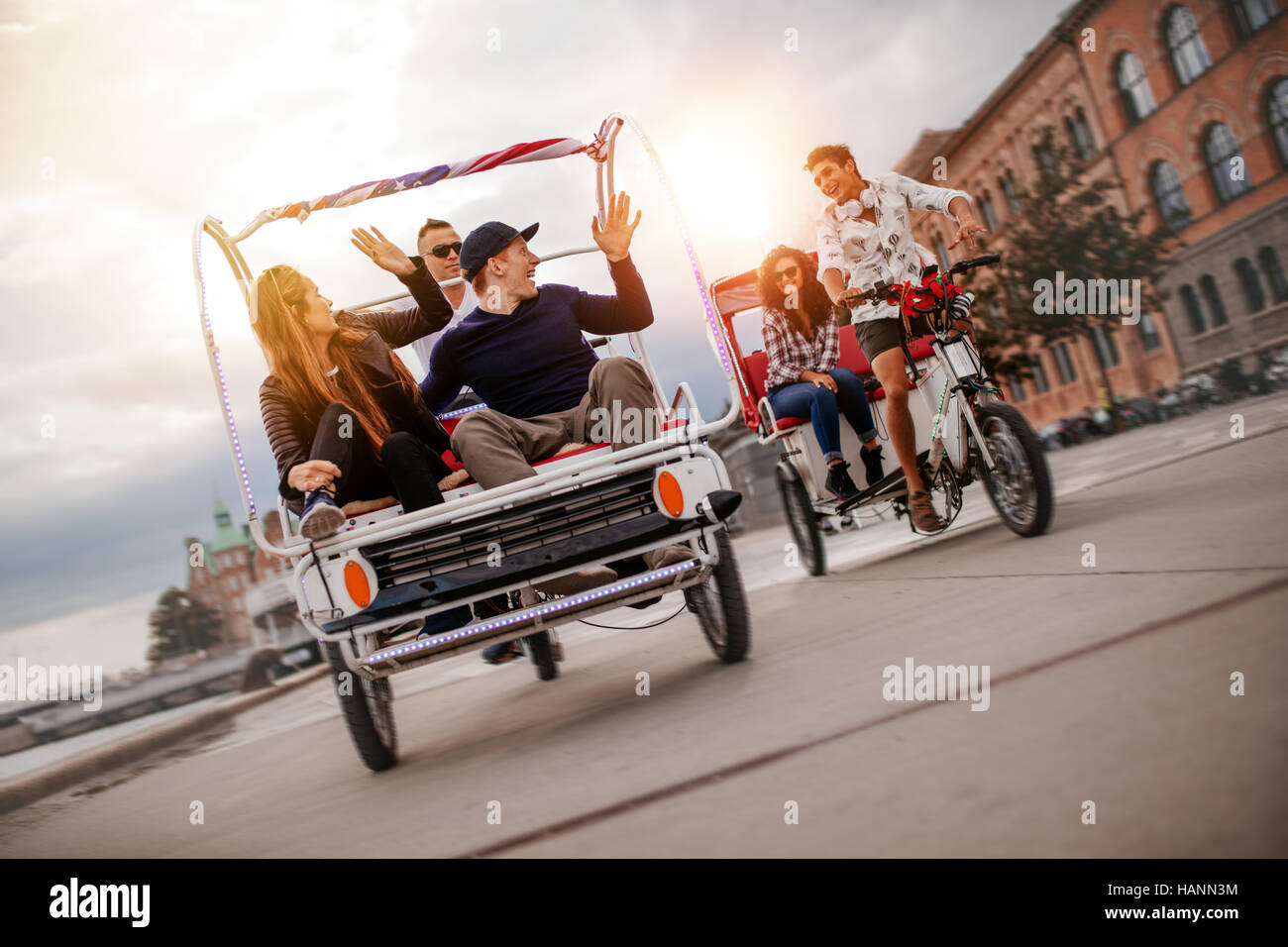 Group of friends having fun with two tricycles on road. Teenagers on tricycle ride on city street. Stock Photo