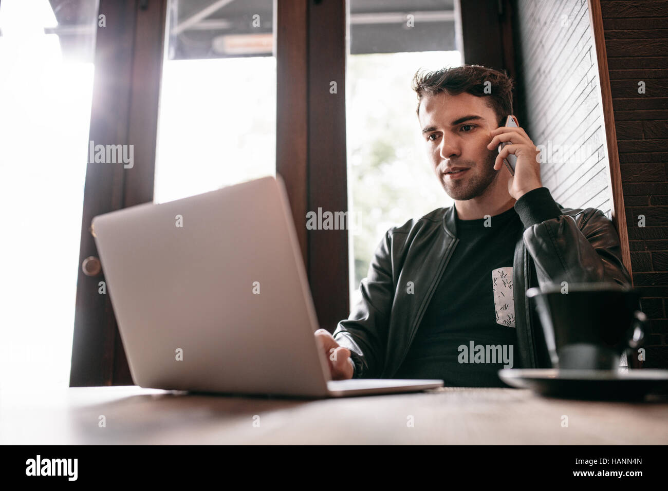 Indoor shot of young man sitting at a cafe table using laptop and talking on mobile phone. Handsome caucasian male sitting at restaurant and working. Stock Photo