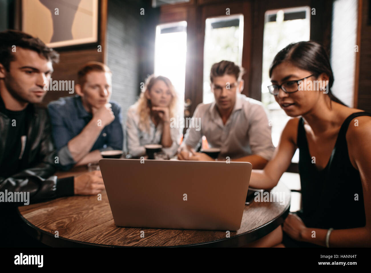 Group of friends sitting around cafe table and looking at laptop. Young men and women at coffee shop using laptop computer. Stock Photo