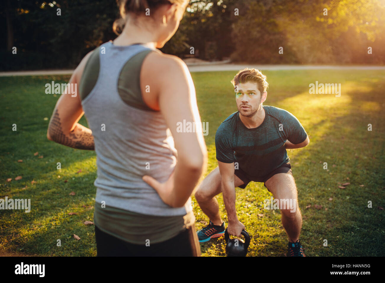 Female trainer guiding man while exercising with kettlebell in park. Personal trainer with man doing weight training in park. Stock Photo
