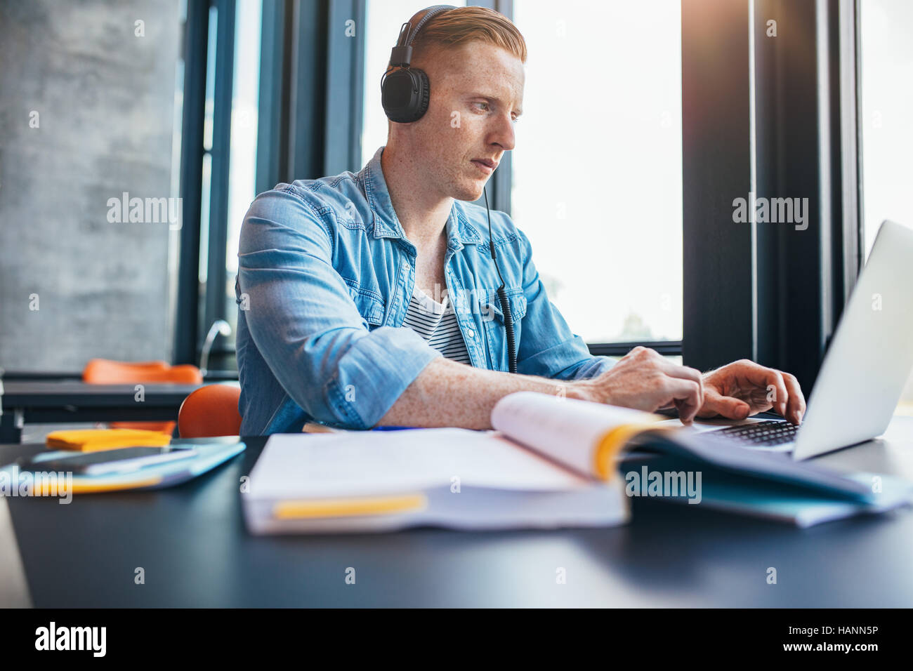 Shot of young caucasian man with headphones studying in library using laptop. University student studying with books and laptop in library. Stock Photo