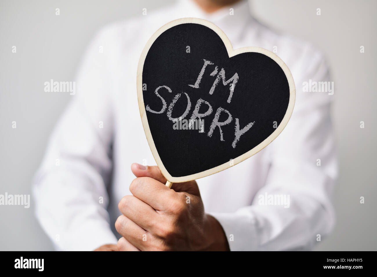 a young caucasian man wearing a white shirt shows a heart-shaped signboard with the text I am sorry written in it Stock Photo
