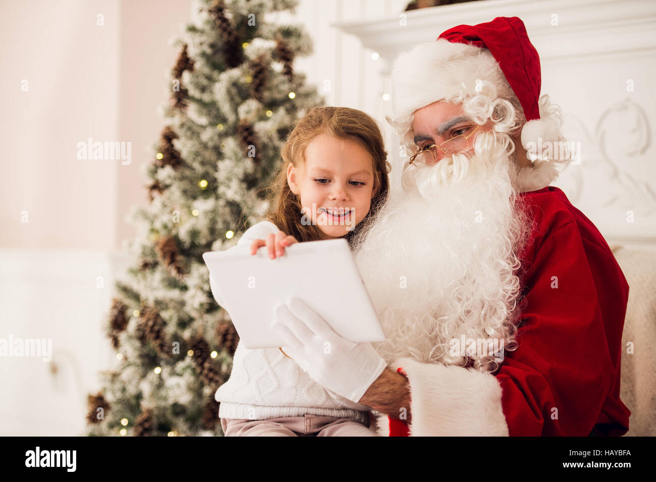 Little girl sitting with Santa using tablet on the armchair at home ...