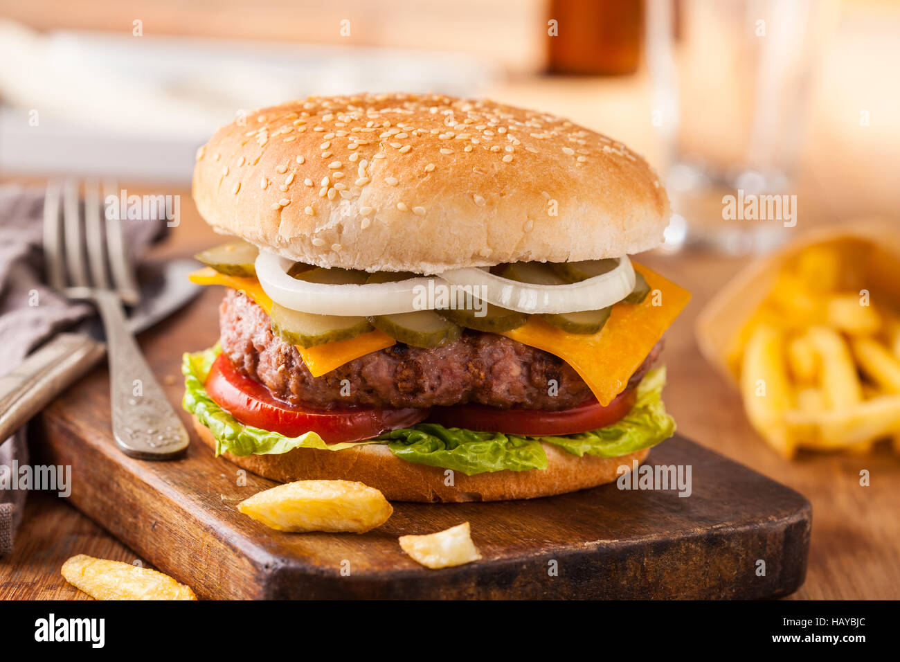 Appetizing hamburger with fries, beer on wooden cutting board. Pickles, onion, and cheddar cheese. Stock Photo