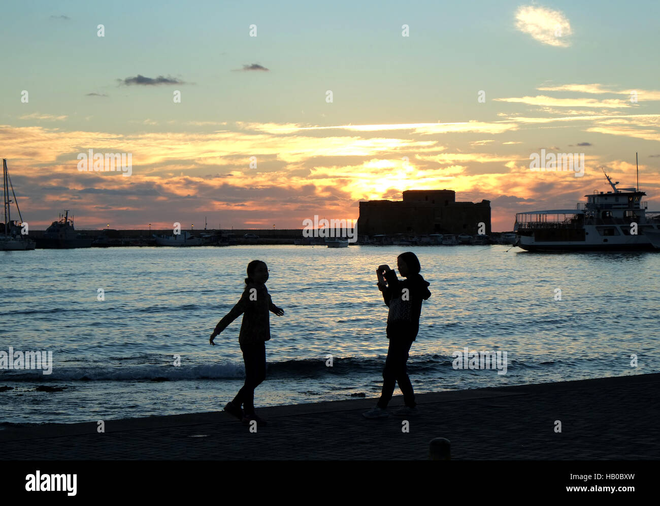 Tourists take pictures on Paphos harbour seafront, with the sun setting behind the medieval fort. Stock Photo