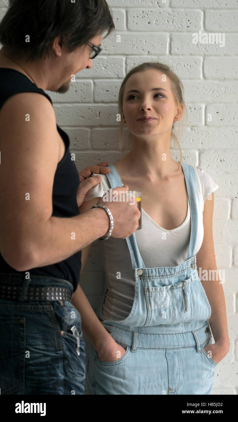 Young man in black singlet gives beauty young caucasian woman in blue denim overalls and a white T-shirt cap of tea front of brick wall Stock Photo