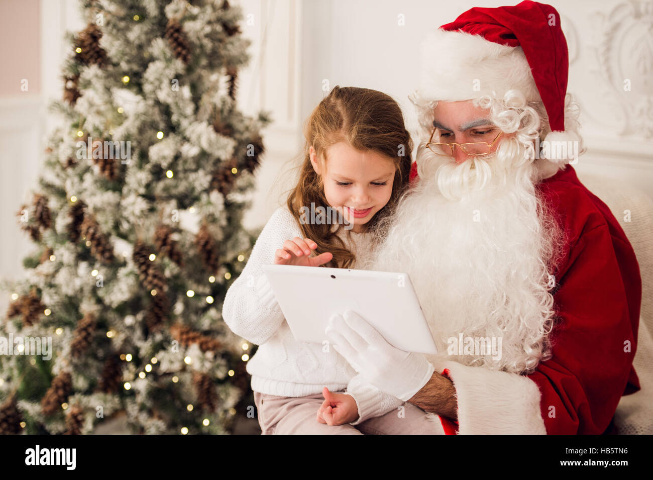 Little girl sitting with Santa using tablet on the armchair at home ...