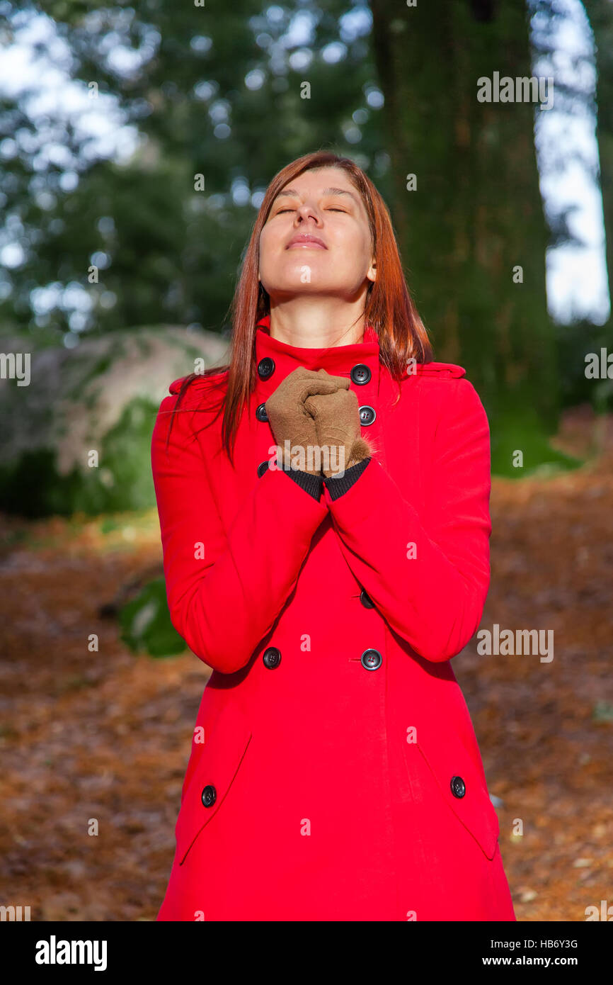 Woman on a forest praying and enjoying the warmth of the winter sunlight, wearing a red long coat Stock Photo