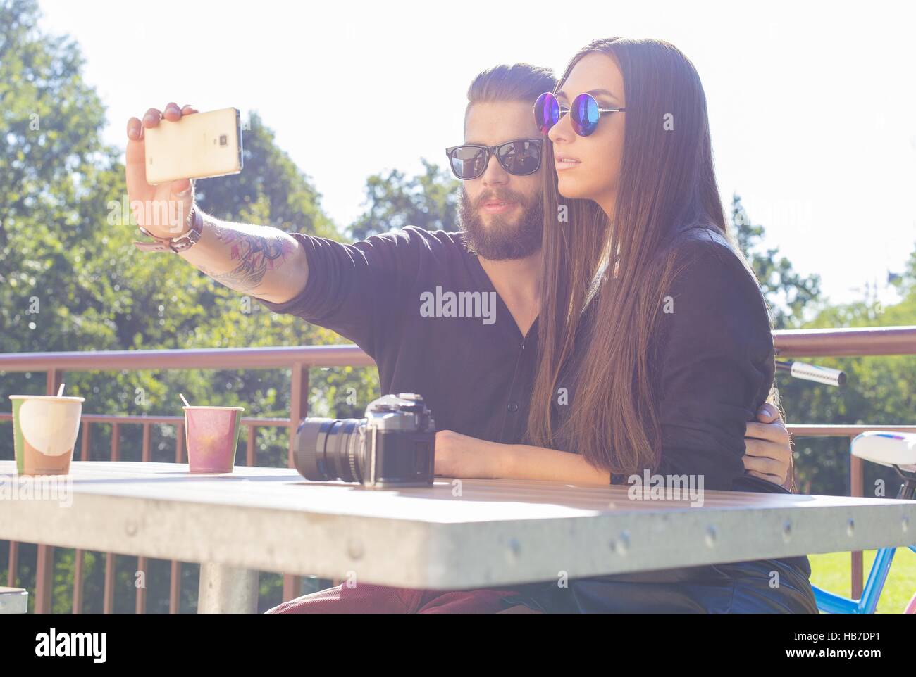 Sitting together at a table in a park and taking selfies. Stock Photo