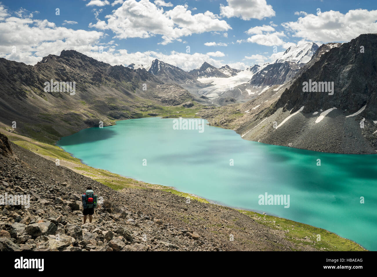 A mountaineer man stops on his trekking trail to observe the snow-capped mountains and Ala Kul Lake, Kyrgyzstan Stock Photo