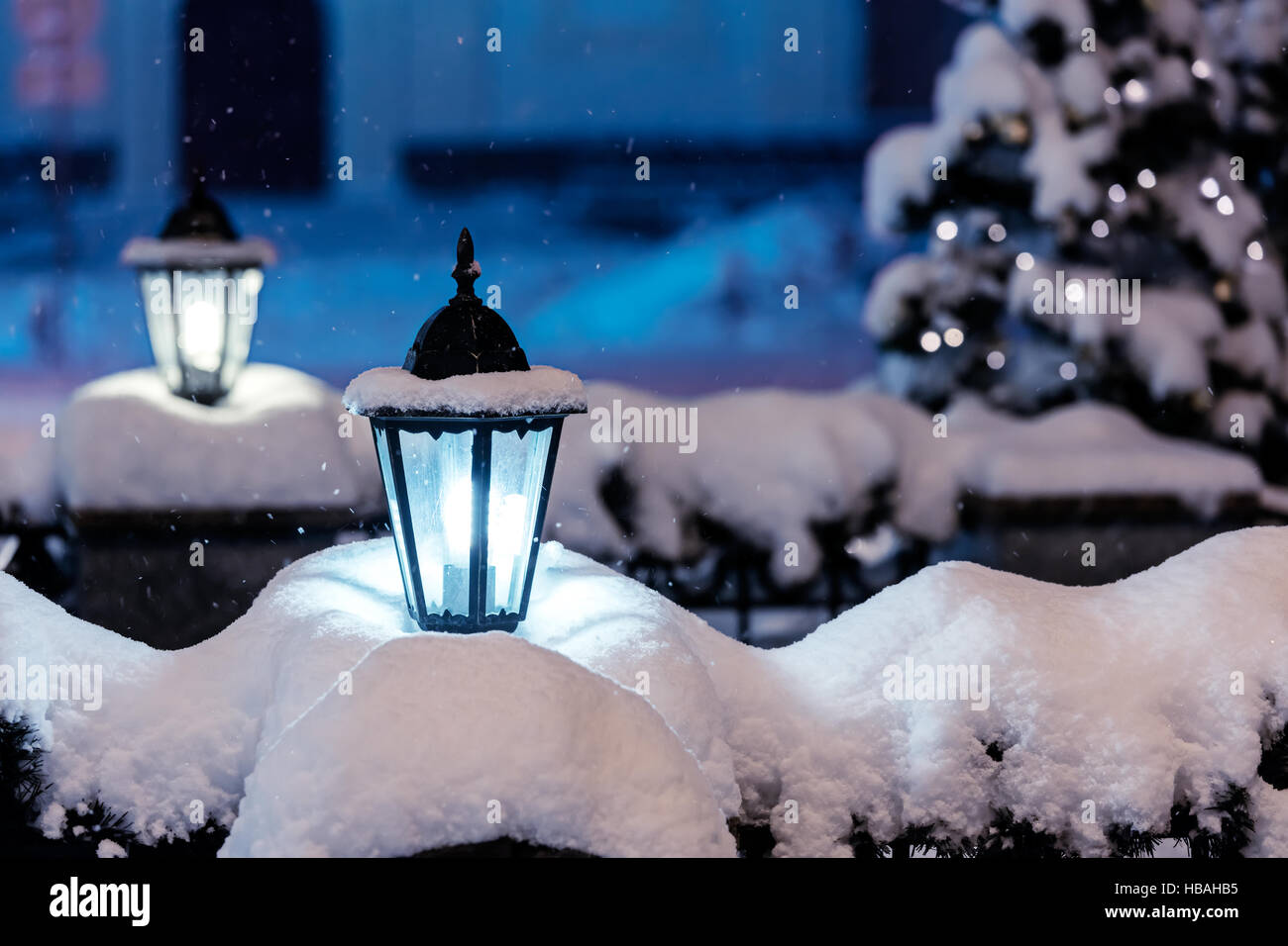 snowy street lamps lit in night city with fir-tree and christmas lights in background Stock Photo