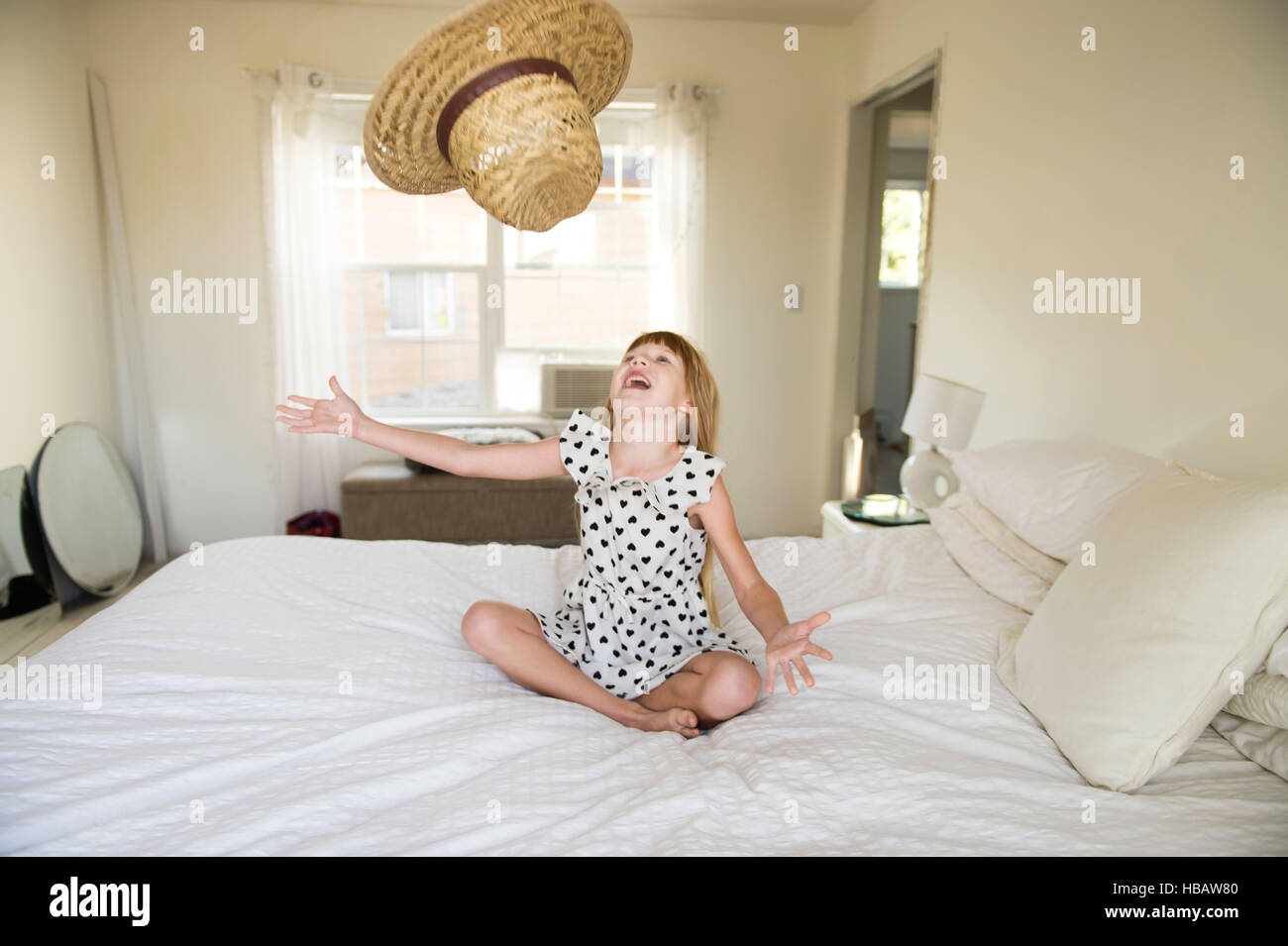 Young girl sitting on bed, throwing straw hat up in air Stock Photo