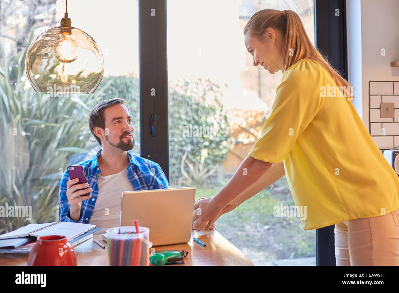 Woman tidying dining table whilst husband using smartphone Stock Photo