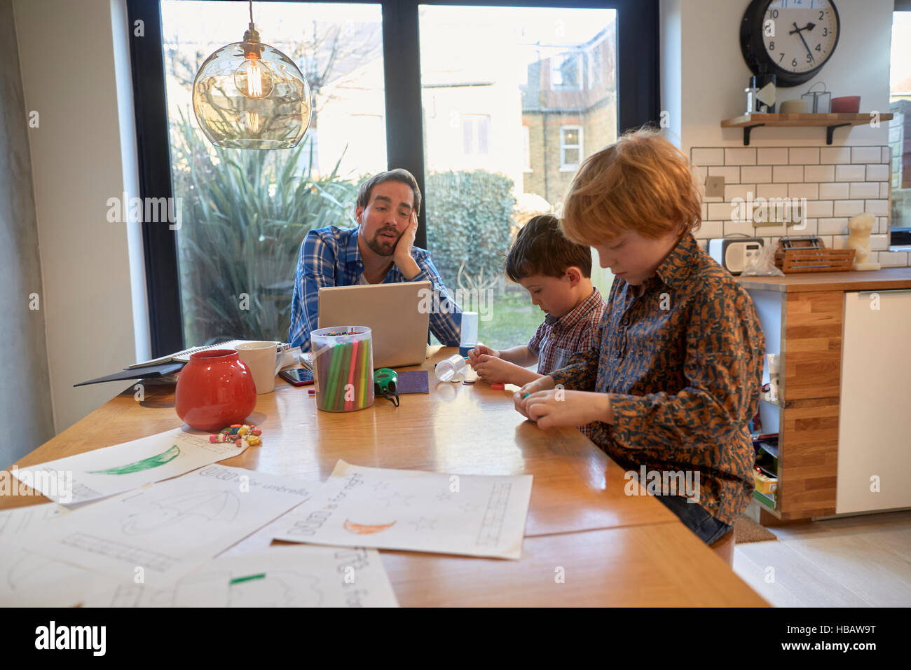 Exhausted man at dining table with laptop whilst son colouring Stock Photo