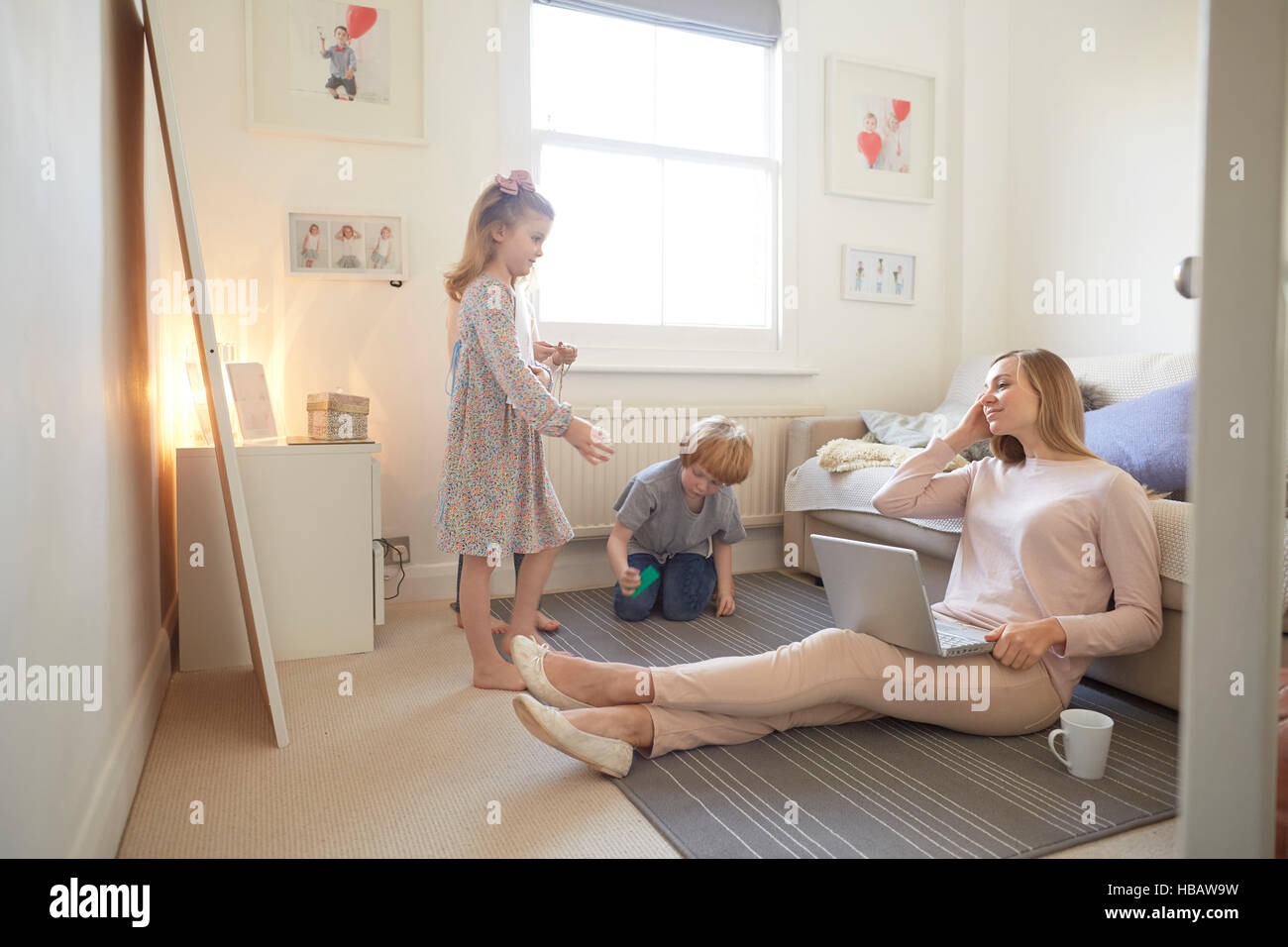 Mid adult woman sitting on living room floor with laptop whilst son and daughter play Stock Photo