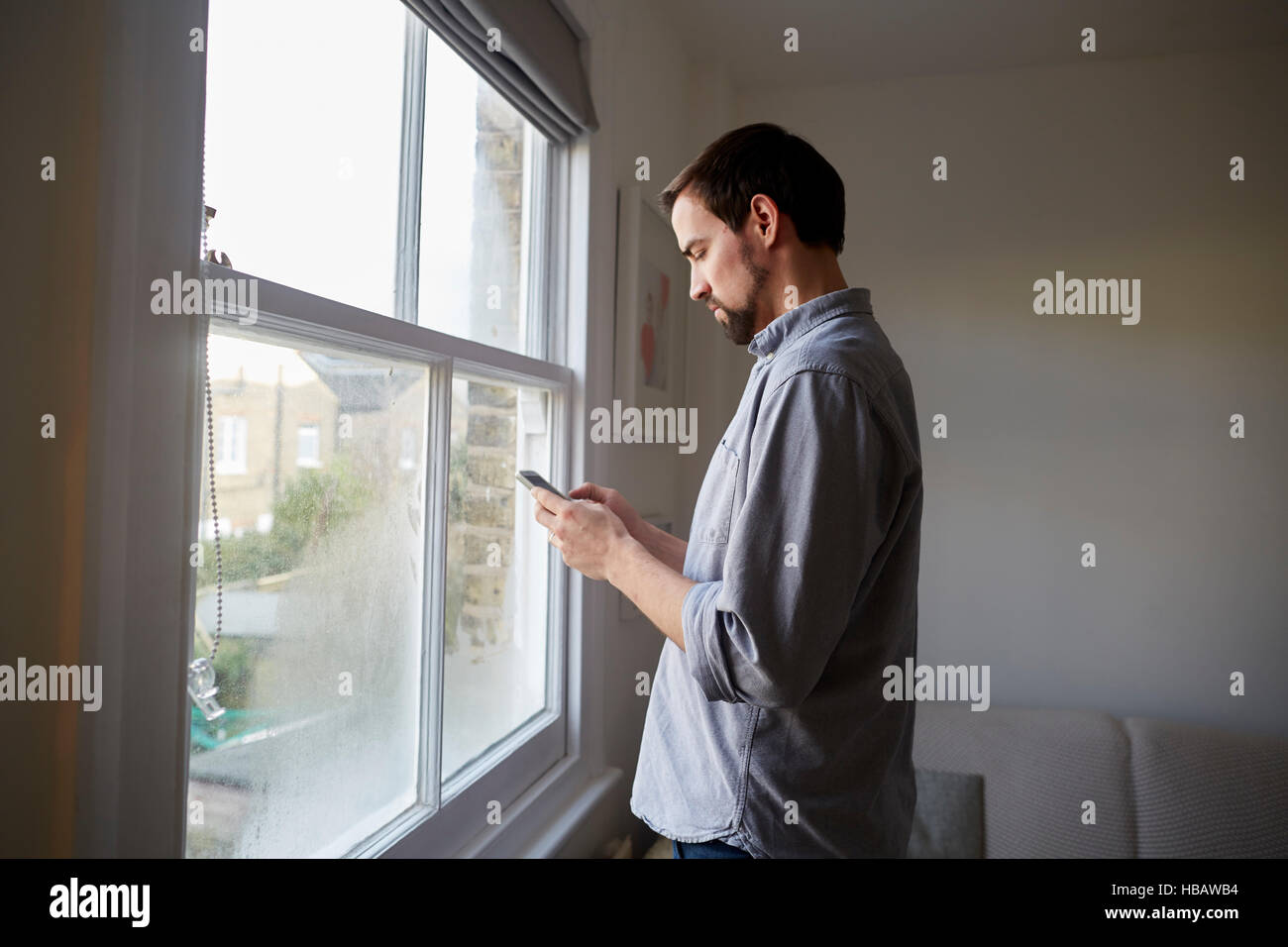 Mid adult man at living room window reading smartphone texts Stock Photo