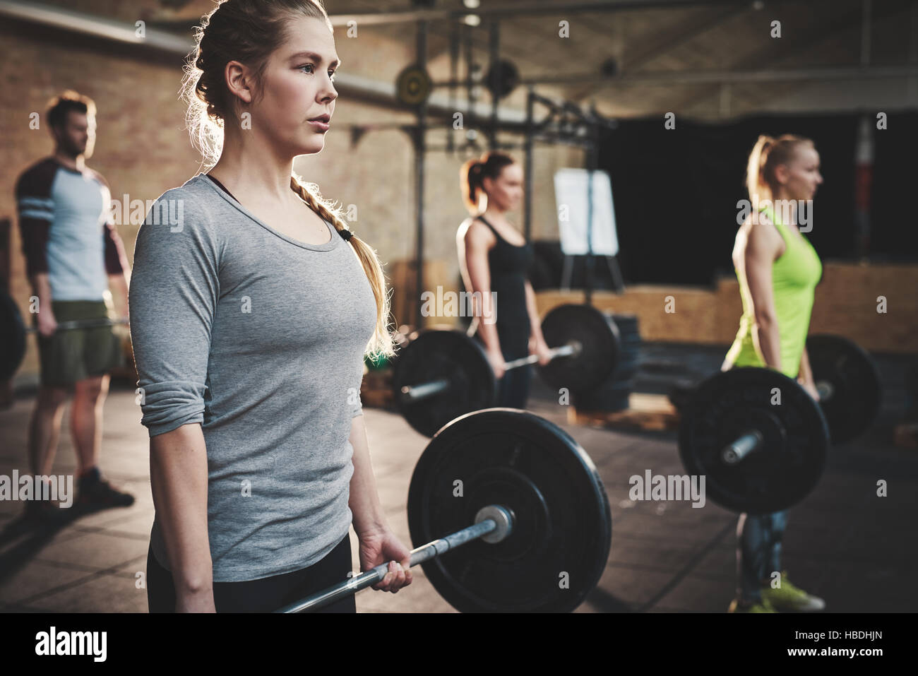 Serious muscular young woman with ponytail and gray shirt performing dead lift barbell exercises with three other trainees Stock Photo