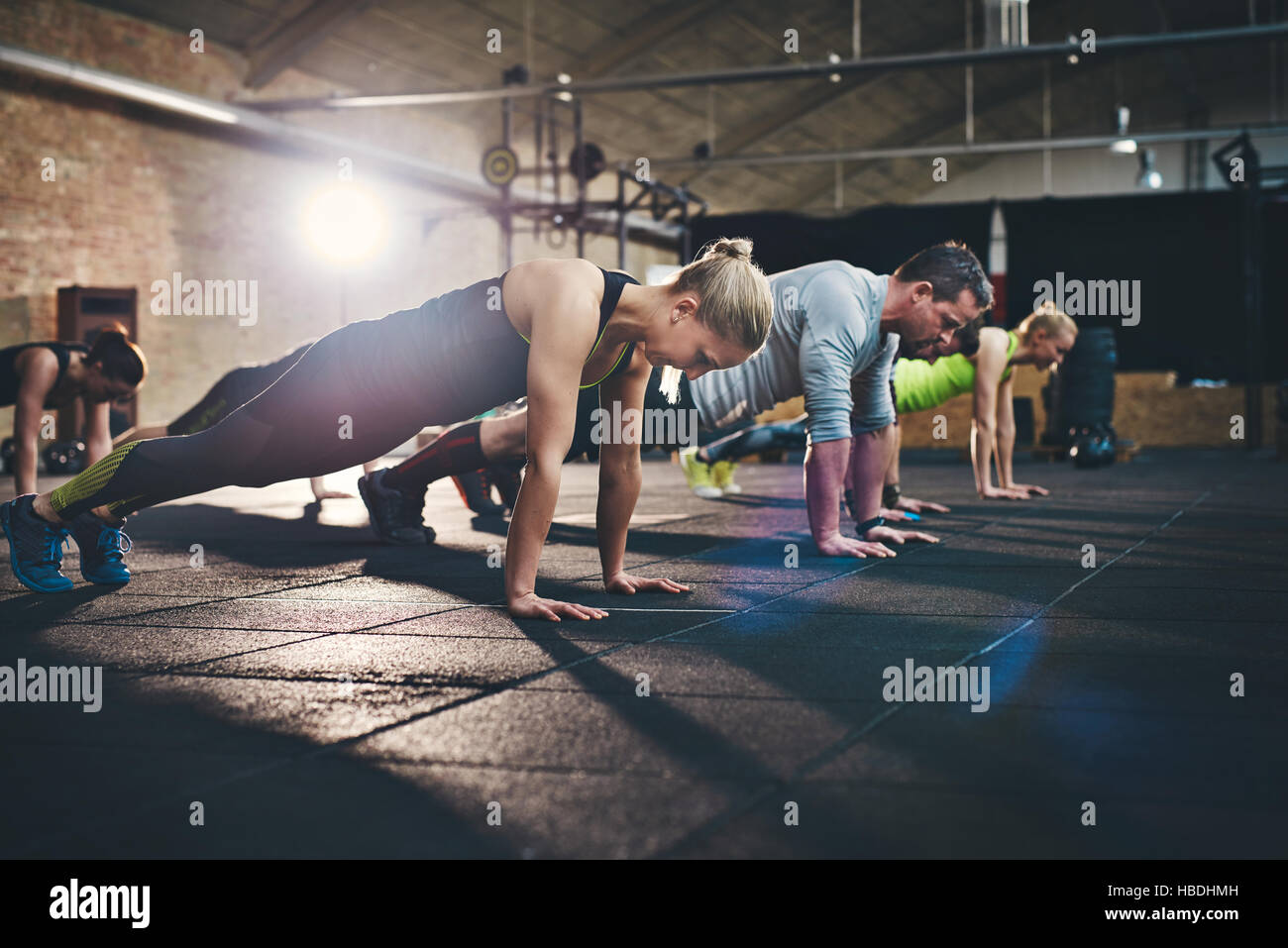 Group of adults doing push up exercises at indoor physical fitness cross-training exercise facility with bright light flare over them Stock Photo