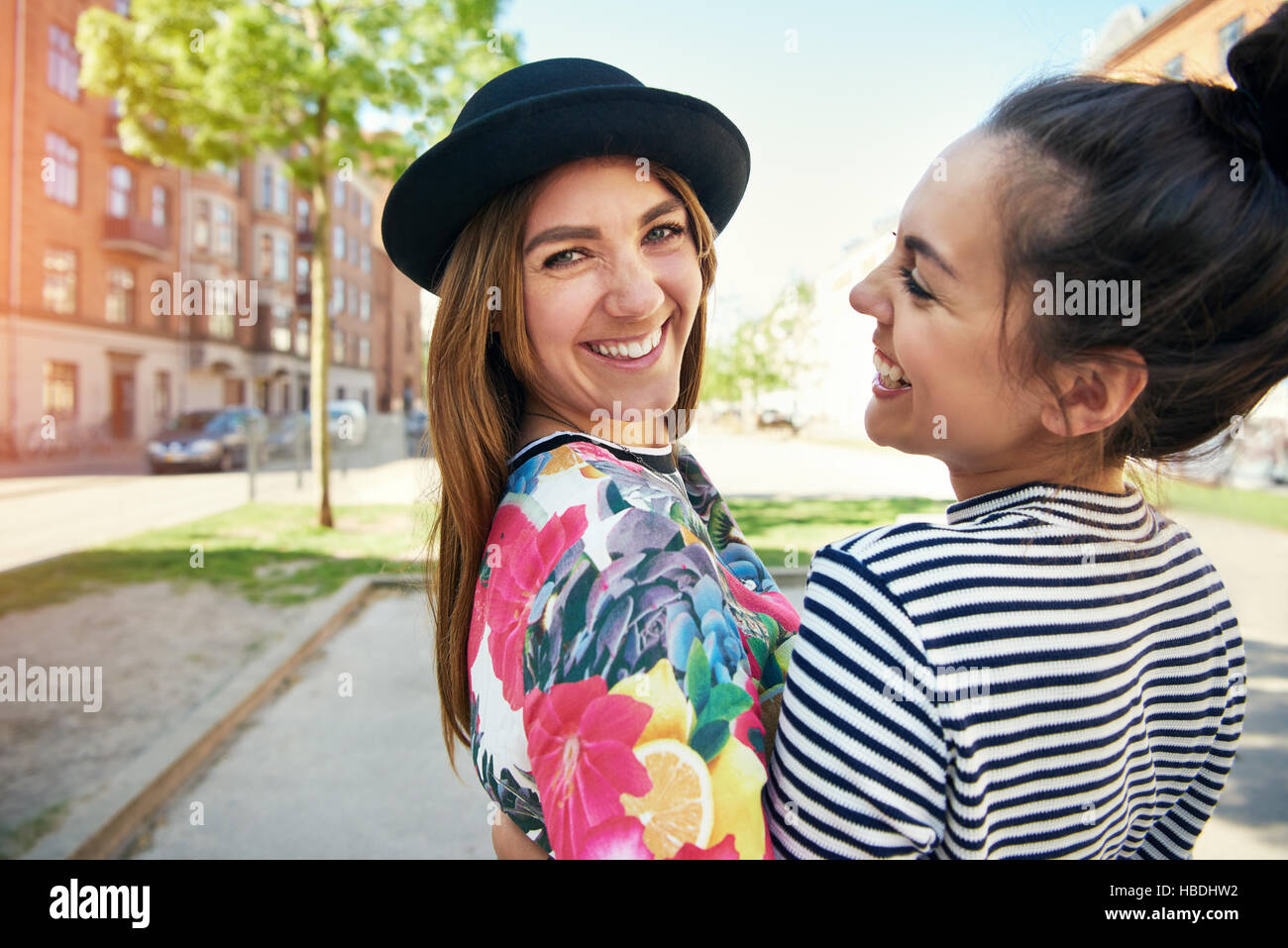 Trendy young woman with a lovely smile wearing a chic little hat standing together with a friend turning to look at the camera in an urban setting Stock Photo