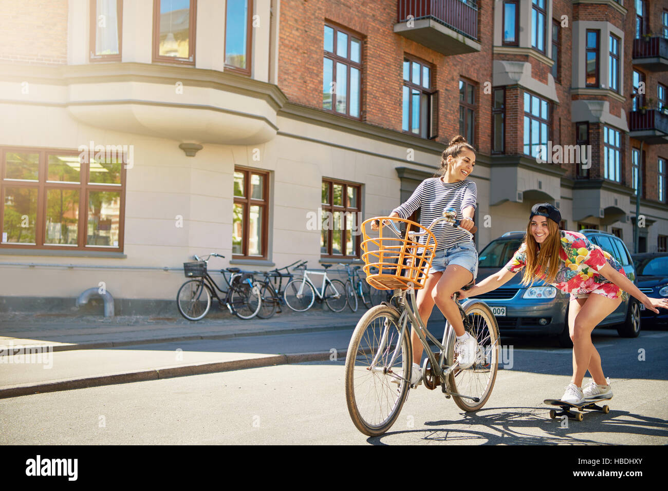 Cute laughing young woman in jeans shorts on bike with basket pulling girl in tie die shirt and black hat holding on while using skateboard Stock Photo