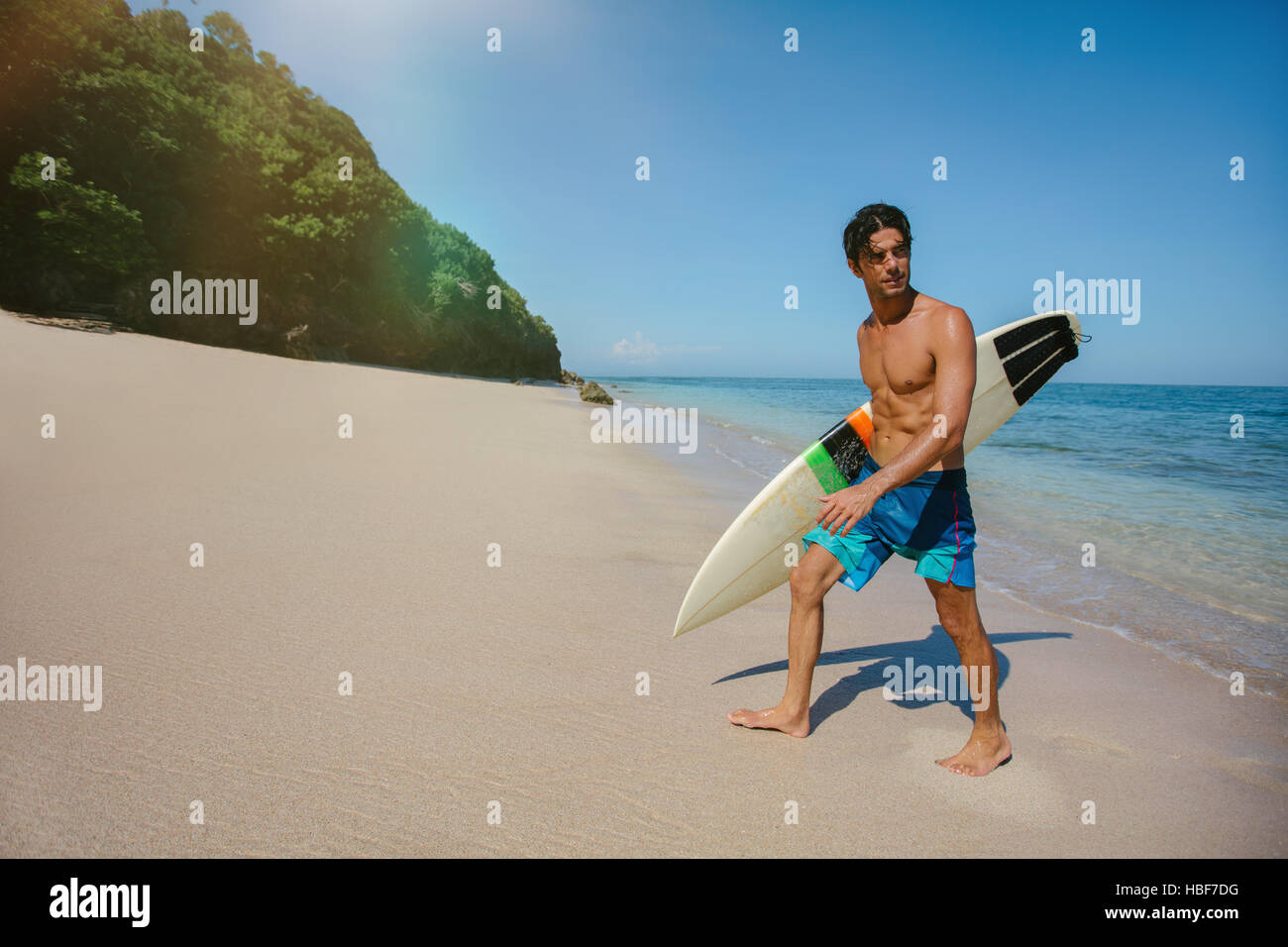 Shot of young male surfer holding surf board walking out of the sea. Caucasian male with surfboard walking along the beach and looking away. Stock Photo