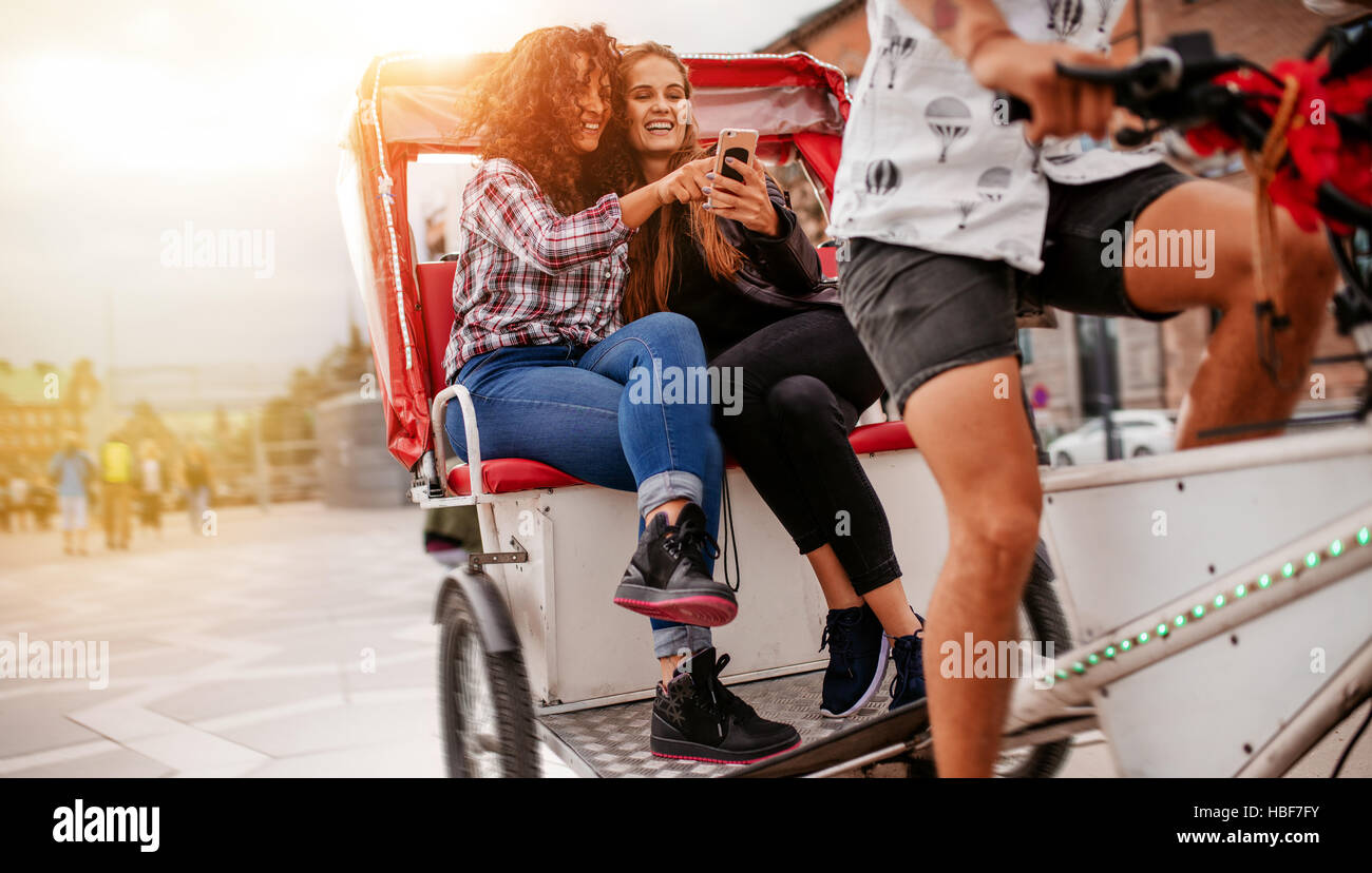 Teenage girls sitting on tricycle using mobile phone. Female friends enjoying tricycle ride on road and using smart phone. Stock Photo