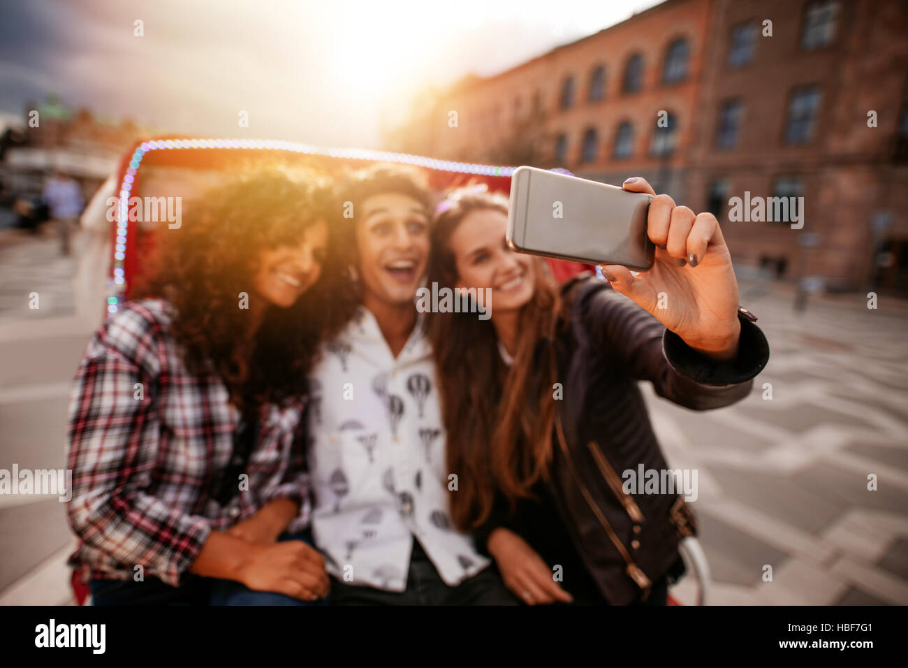 Three young friends taking selfie on tricycle ride. Group of people riding on tricycle bike and taking self portrait. Focus on mobile phone in female Stock Photo