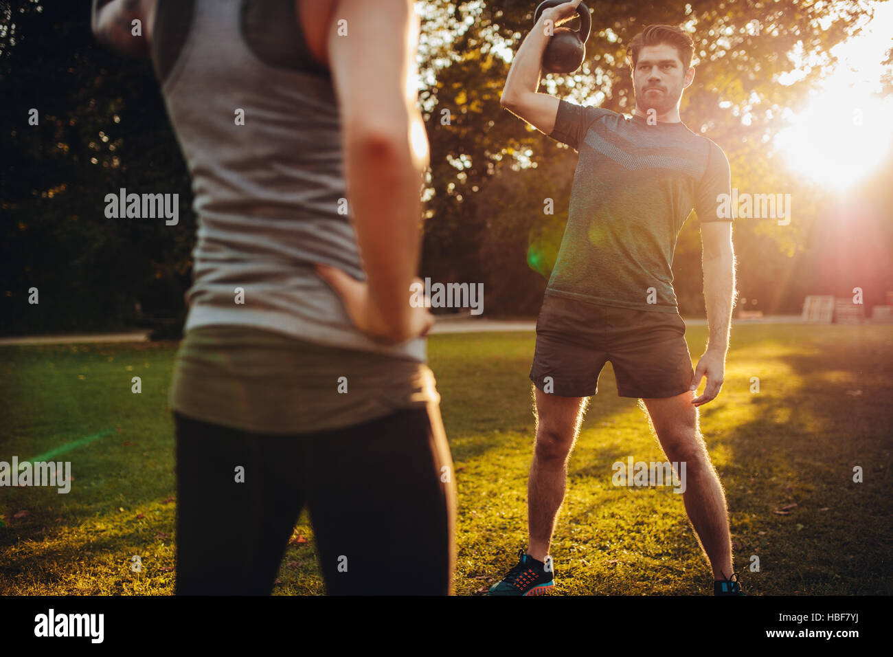 Fit young man training with kettlebell in the park. Healthy man exercising with female trainer in front. Stock Photo