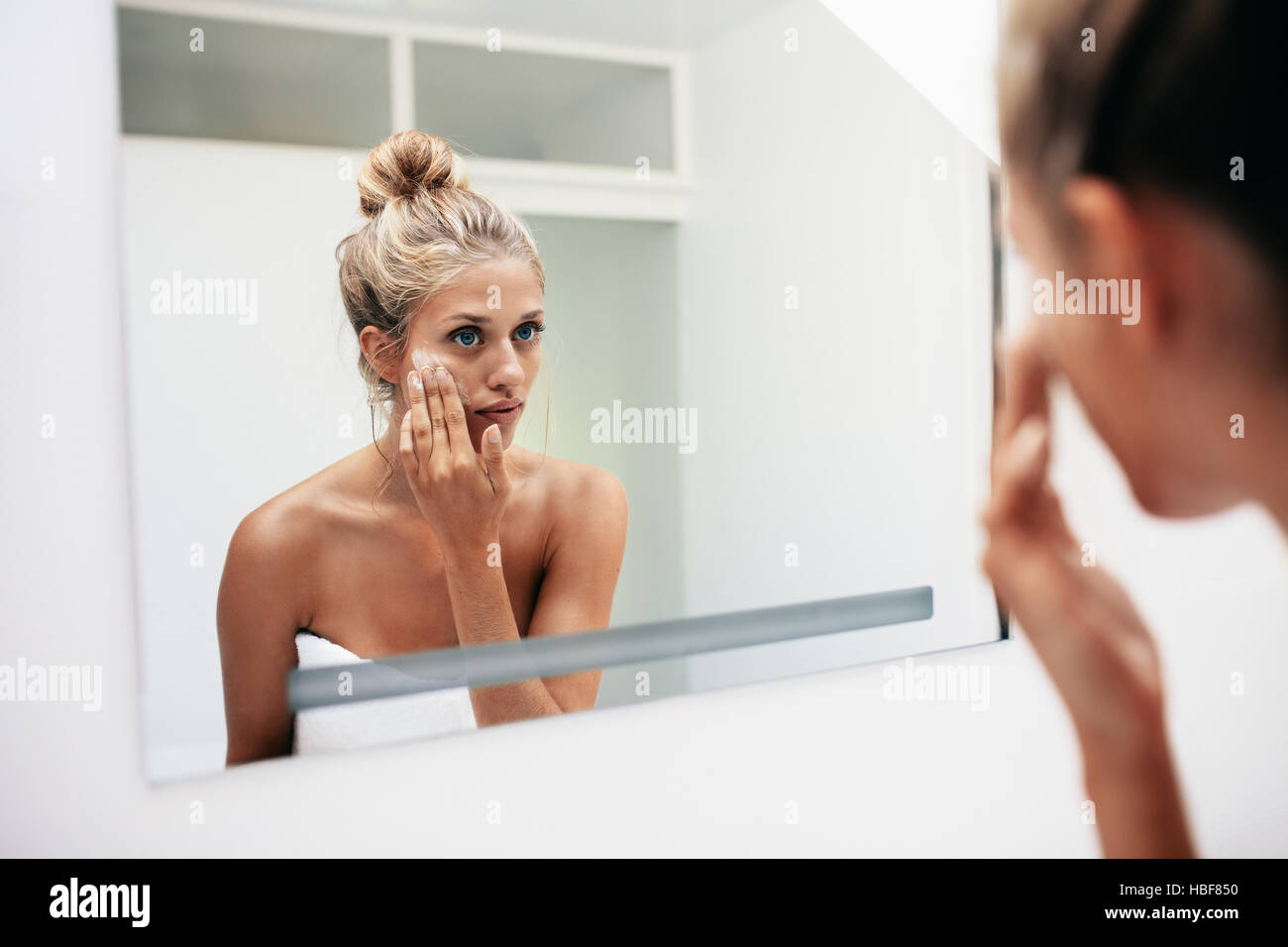 Reflection of a female in mirror rubbing cosmetic cream on her face. Female putting on moisturizer on her facial skin in bathroom. Stock Photo