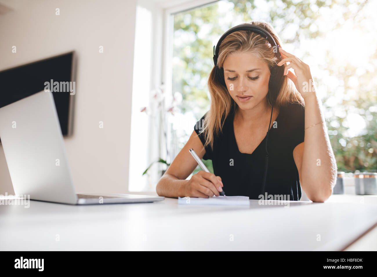 Indoor shot of woman studying in kitchen. Female wearing headphones and writing with laptop on table. Stock Photo