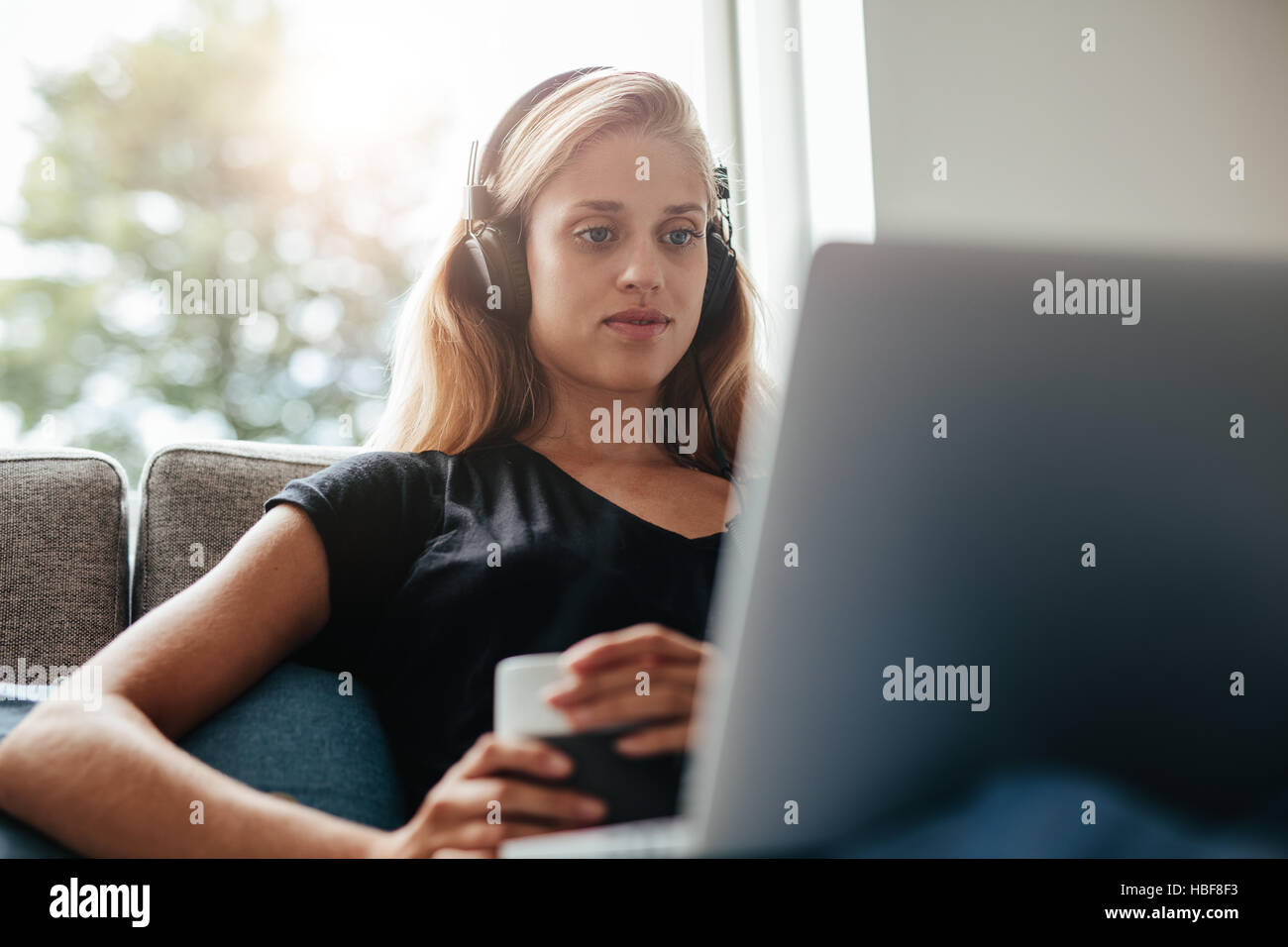 Young woman with headphones looking at laptop. She is sitting on couch in living room at home. Stock Photo