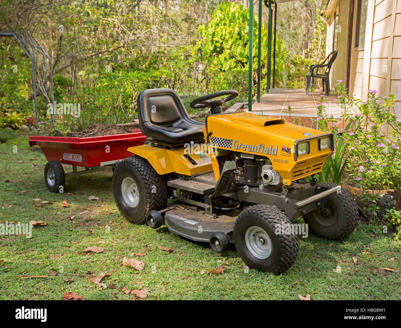 Yellow ride-on mower towing small red trailer full of soil on lawn beside house with lush garden of shrubs and trees Stock Photo