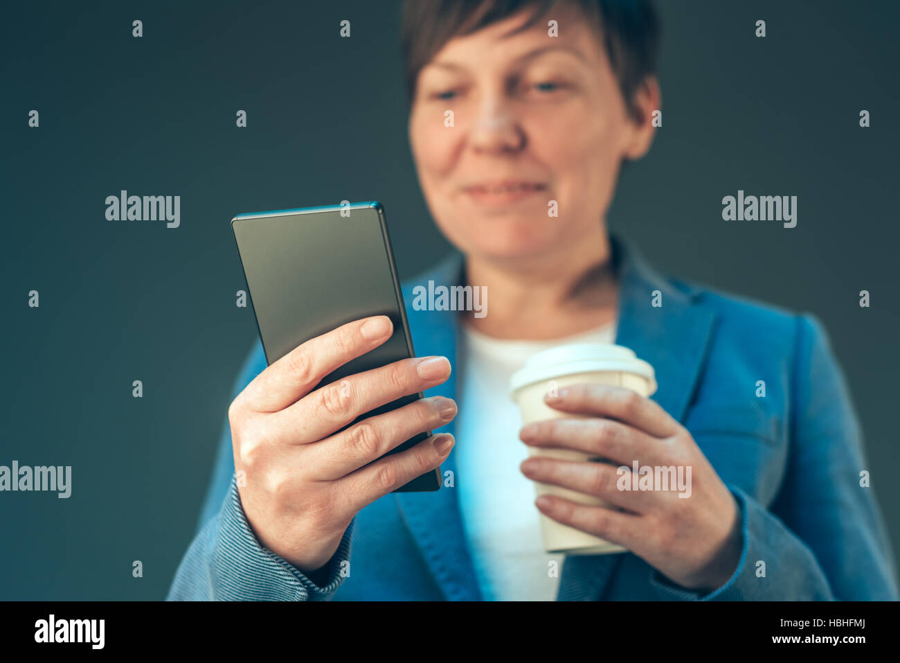 Satisfied young adult businesswoman drinking coffee to go and looking at the mobile phone Stock Photo