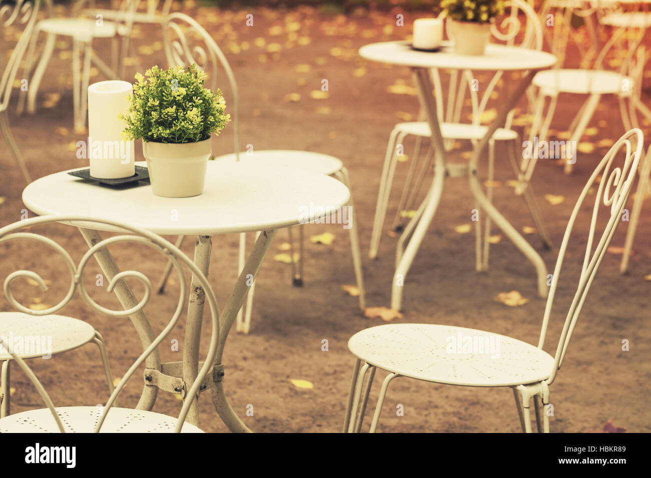Outdoor cafeteria background interior, metal white chairs and tables with decorative green plants in pots and candles, vintage stylized photo, warm to Stock Photo