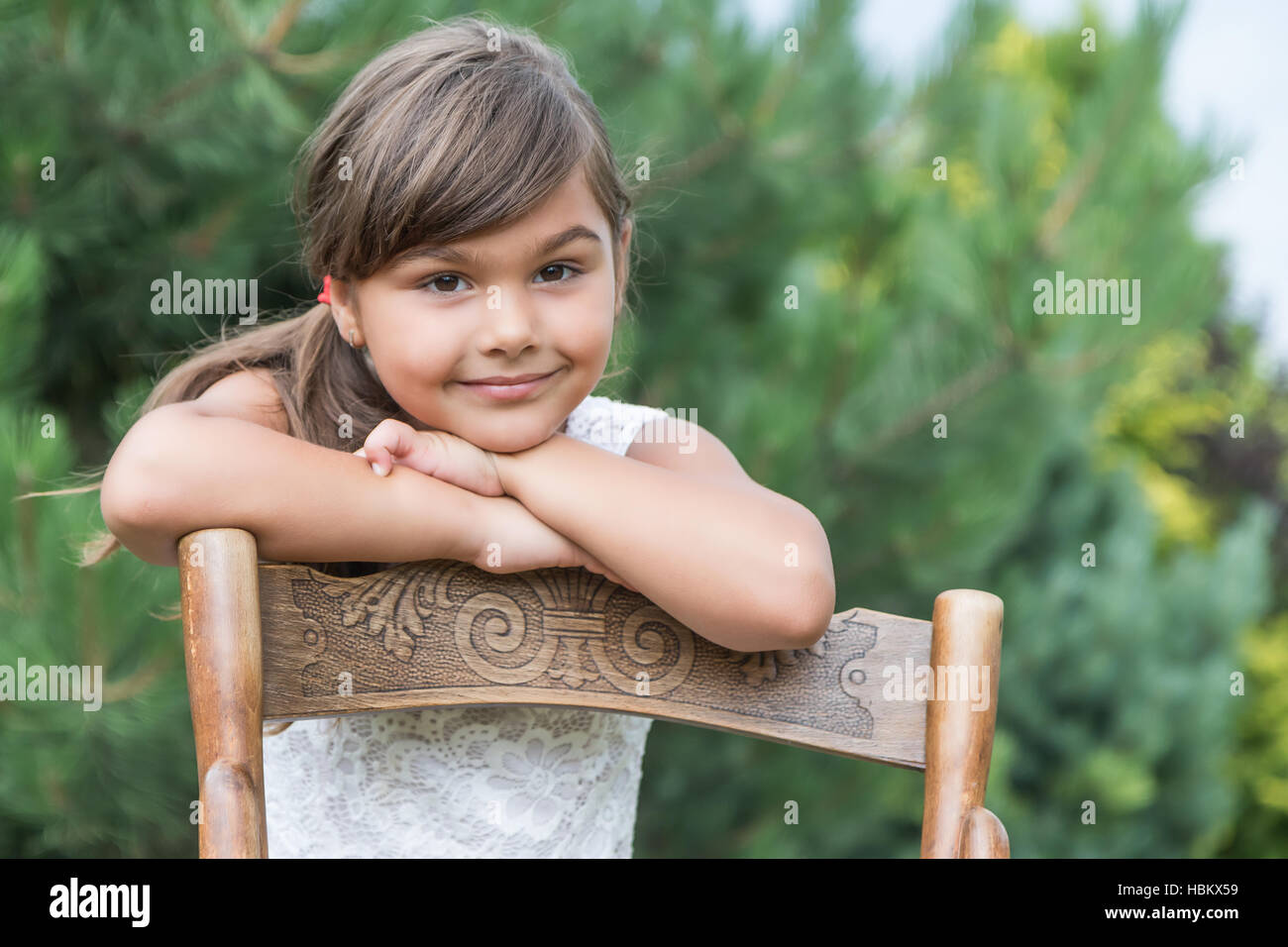 Portrait of cool long haired  brunette little girl leaning on an vintage wooden chair outdoors. Stock Photo