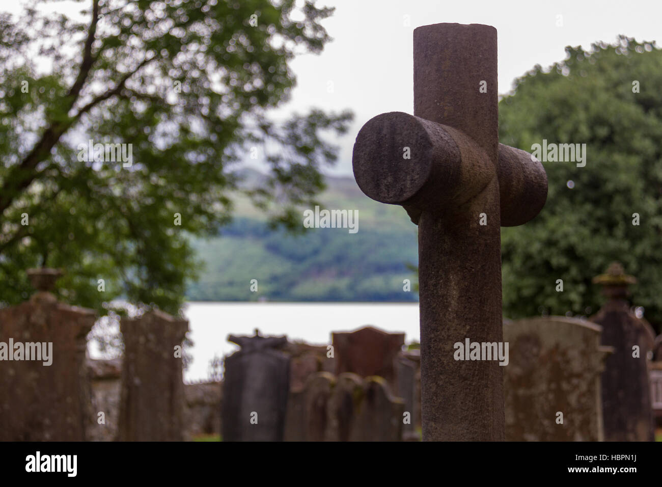 Cross and tombstones in cemetery with lake in background, in Luss parish church yard in Scotland Stock Photo