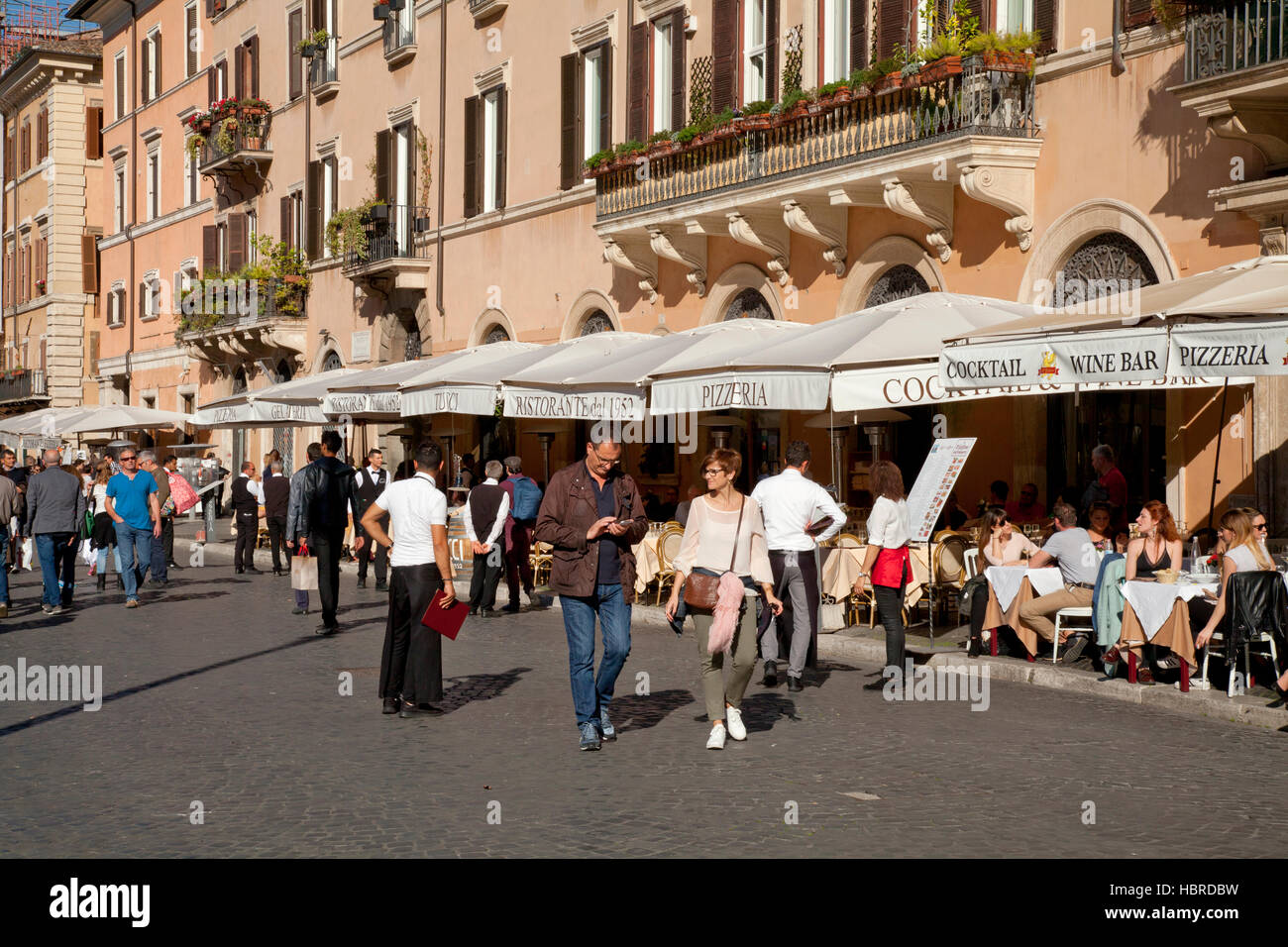 Piazza Navona, Rome, tourists dining at restaurant al fresco in the sunshine, with waiters and couple walking by Stock Photo