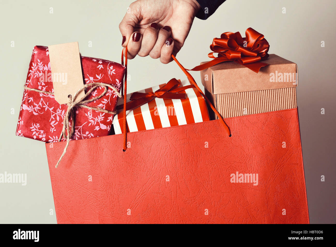 closeup of the hand of a young caucasian woman with her fingernails painted red holding a red shopping bag full of gifts wrapped in different papers Stock Photo