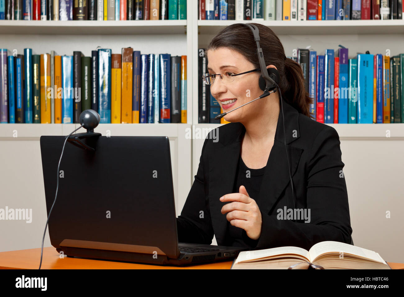 Mature woman with headset, laptop and webcam in front of a lot of books,  giving online legal advice via a video call Stock Photo - Alamy
