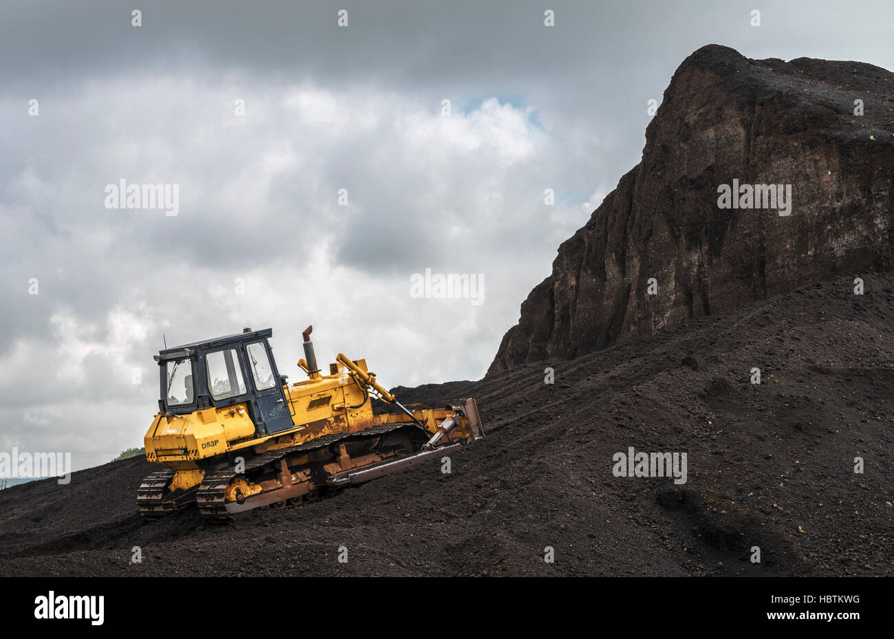 A yellow bulldozer at the foot of a giant pile of peat Stock Photo