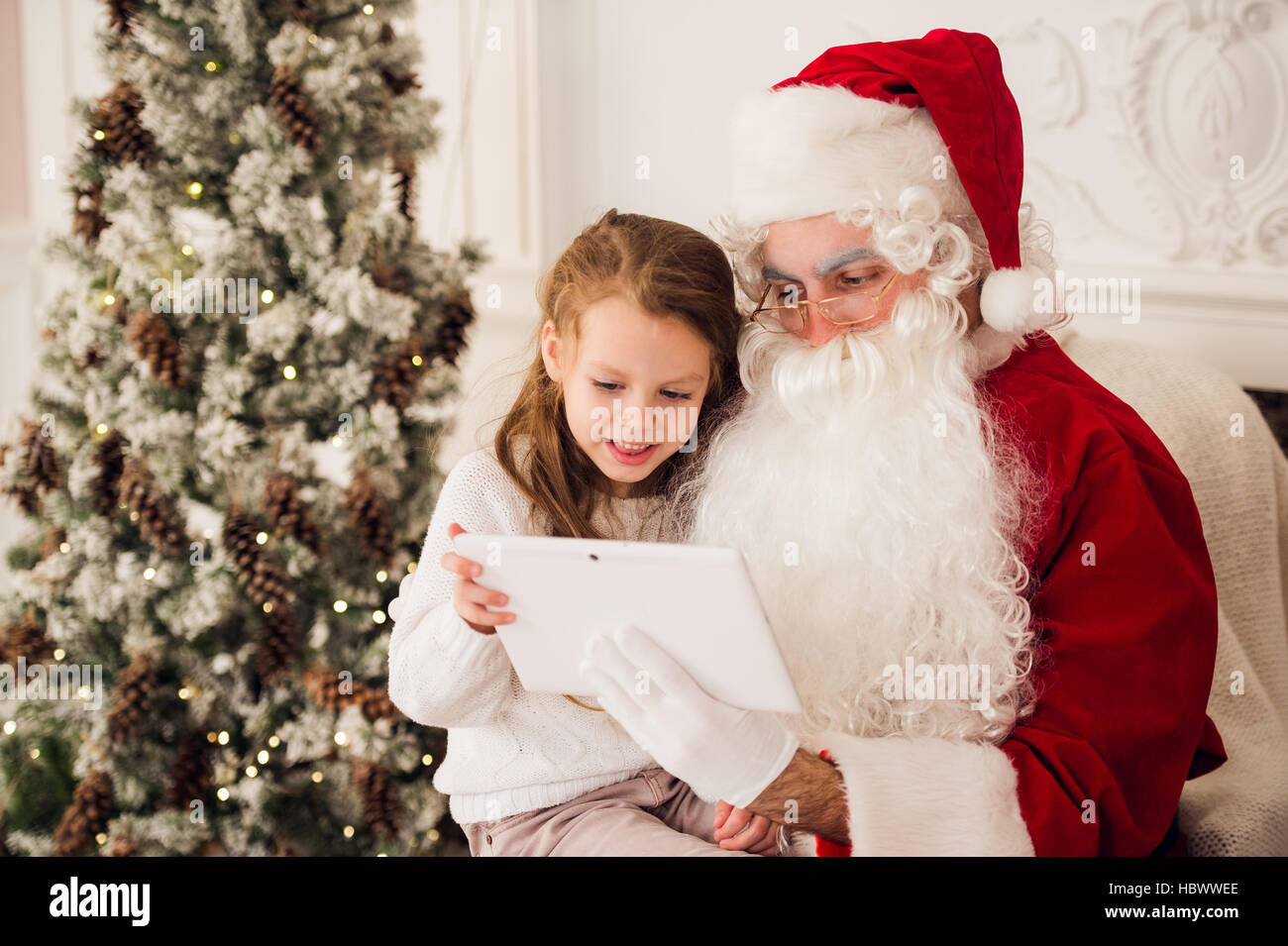 Little girl sitting with Santa using tablet on the armchair at home living room Stock Photo