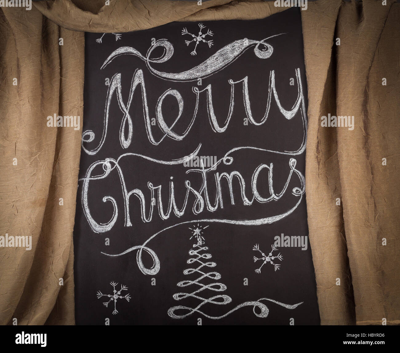 Merry Christmas hand drawn lettering with chalk on blackboard framed by golden curtains. Elegant unique design with snowflakes and Christmas tree Stock Photo