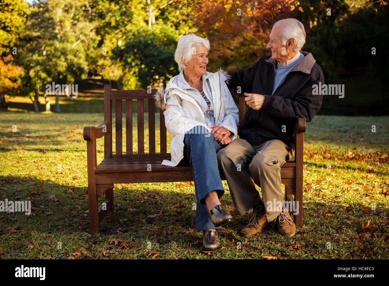 Elderly couple sitting on bench smiling at each other in park Stock Photo