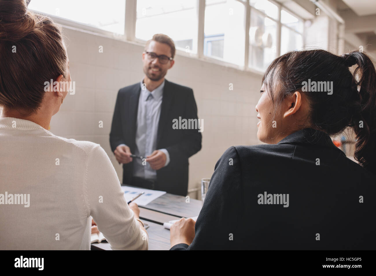 Two young woman sitting in a meeting in office, with businessman standing in background. Business presentation in meeting room. Stock Photo