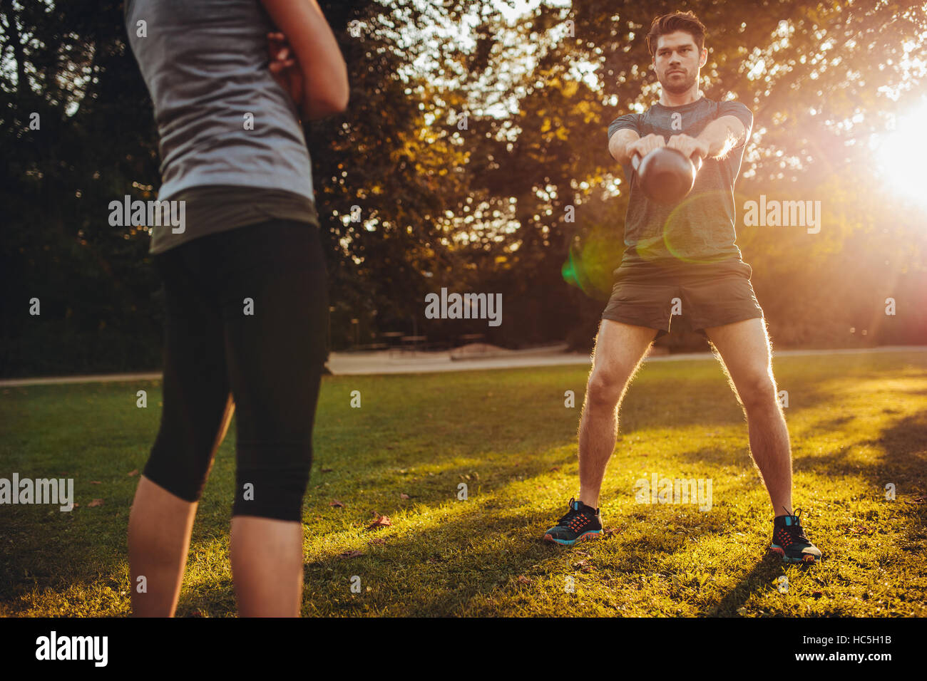 Young man doing kettlebell weight workout with personal female trainer in the park. Fit man swinging kettle bell with fitness instructor. Stock Photo