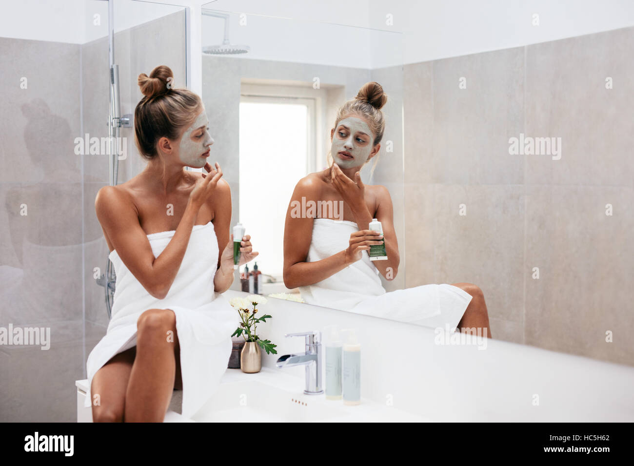 Young woman applying facial mud clay mask to her face in bathroom. Beautiful female wrapped in towel looking into mirror and doing facial beauty treat Stock Photo
