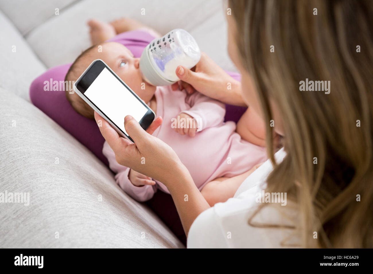Mother using mobile phone while feeding her baby with milk bottle Stock Photo