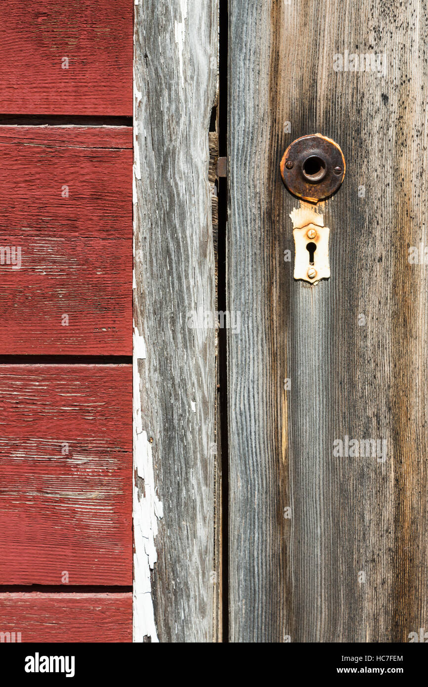 Close up of door handle broken off, rusty metal and old white colored lock old worn wood door and red side wood building. Stock Photo